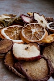 A close-up view of various dried fruit slices, including oranges, apples, and others. The texture of the dried fruits is rugged with visible details of the fruit's surface and rind. The lighting creates a warm and earthy tone.