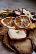 A close-up view of various dried fruit slices, including oranges, apples, and others. The texture of the dried fruits is rugged with visible details of the fruit's surface and rind. The lighting creates a warm and earthy tone.