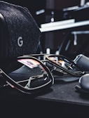 A pair of reflective sunglasses are placed on a dark, soft-textured surface. Nearby, there is a grey, fabric-covered virtual reality headset with a distinctive 'G' logo visible. The reflections in the sunglasses show hints of colorful lights from the surroundings.