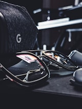 A pair of reflective sunglasses are placed on a dark, soft-textured surface. Nearby, there is a grey, fabric-covered virtual reality headset with a distinctive 'G' logo visible. The reflections in the sunglasses show hints of colorful lights from the surroundings.