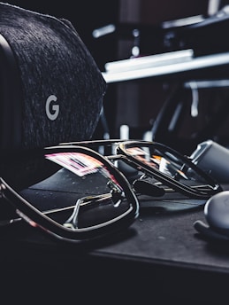 A pair of reflective sunglasses are placed on a dark, soft-textured surface. Nearby, there is a grey, fabric-covered virtual reality headset with a distinctive 'G' logo visible. The reflections in the sunglasses show hints of colorful lights from the surroundings.