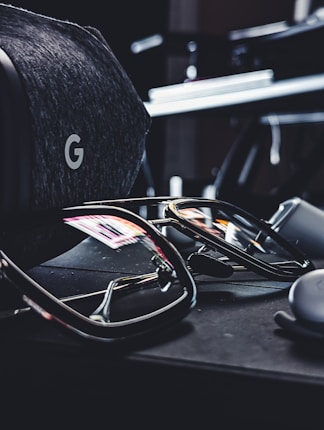 A pair of reflective sunglasses are placed on a dark, soft-textured surface. Nearby, there is a grey, fabric-covered virtual reality headset with a distinctive 'G' logo visible. The reflections in the sunglasses show hints of colorful lights from the surroundings.