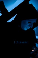 Close-up of a menacing masked villain holding a glowing weapon in a dimly lit alley.