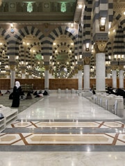 The interior of a large mosque with grand arches and columns, featuring intricate patterns and designs. The floor is made of polished marble, and several people are seen sitting or walking. The lighting is warm, with numerous lantern-style lamps hanging from the ceiling.