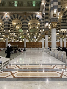 The interior of a large mosque with grand arches and columns, featuring intricate patterns and designs. The floor is made of polished marble, and several people are seen sitting or walking. The lighting is warm, with numerous lantern-style lamps hanging from the ceiling.