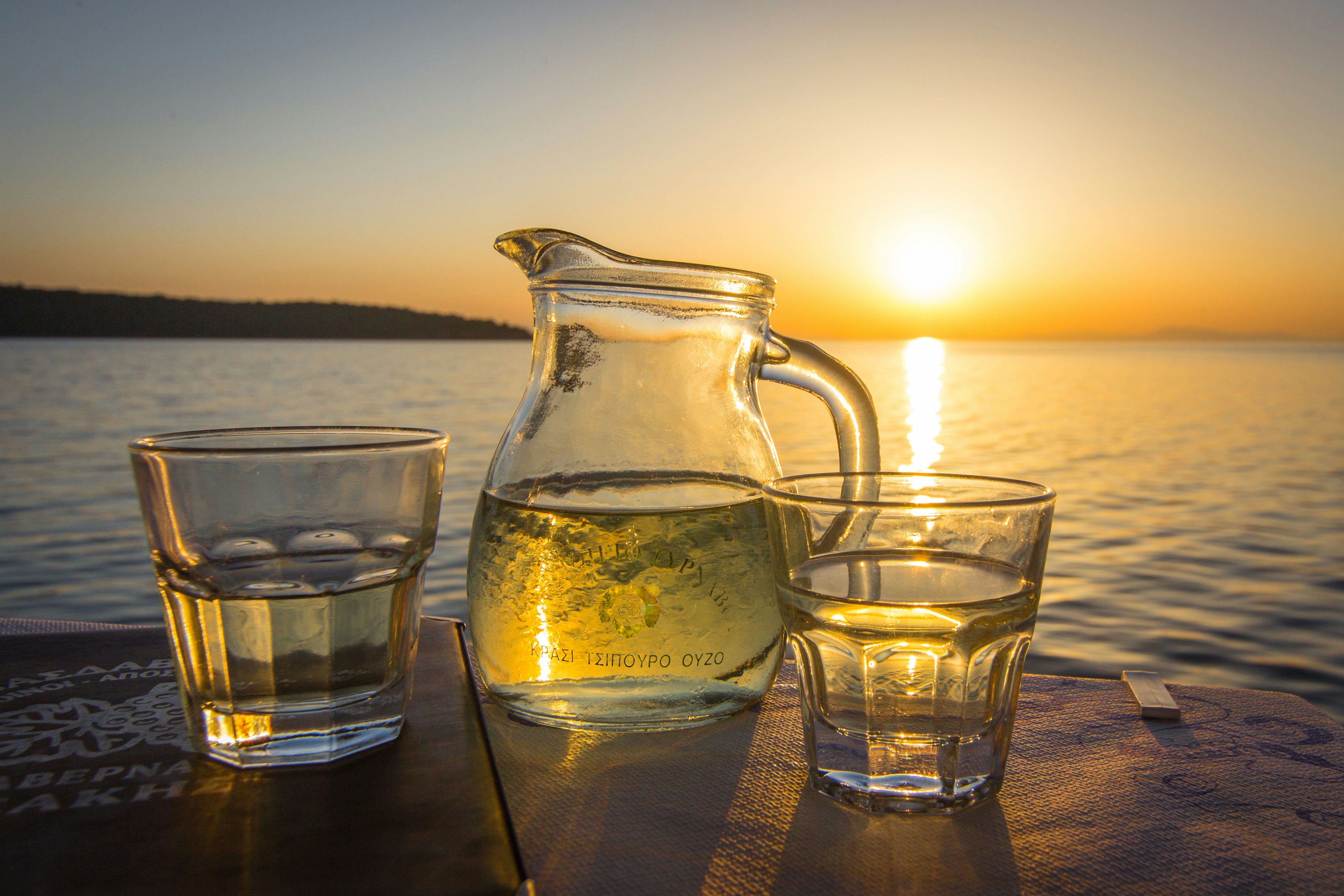 a pitcher of water and two glasses on a table, A jug of Greek house wine in the sunset