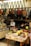 The image depicts the interior of a small shop selling various types of bags and pouches. Multiple handbags, predominantly in earthy tones, are hanging from hooks above the counter. The counter is cluttered with items, and a middle-aged man wearing a face mask and white shirt is seen reading a book behind the counter. In the foreground, there are baskets with different wallets, pouches, and boxes, each marked with price tags in yen.