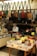 The image depicts the interior of a small shop selling various types of bags and pouches. Multiple handbags, predominantly in earthy tones, are hanging from hooks above the counter. The counter is cluttered with items, and a middle-aged man wearing a face mask and white shirt is seen reading a book behind the counter. In the foreground, there are baskets with different wallets, pouches, and boxes, each marked with price tags in yen.