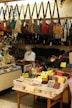 The image depicts the interior of a small shop selling various types of bags and pouches. Multiple handbags, predominantly in earthy tones, are hanging from hooks above the counter. The counter is cluttered with items, and a middle-aged man wearing a face mask and white shirt is seen reading a book behind the counter. In the foreground, there are baskets with different wallets, pouches, and boxes, each marked with price tags in yen.