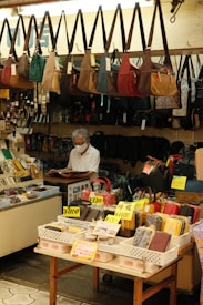The image depicts the interior of a small shop selling various types of bags and pouches. Multiple handbags, predominantly in earthy tones, are hanging from hooks above the counter. The counter is cluttered with items, and a middle-aged man wearing a face mask and white shirt is seen reading a book behind the counter. In the foreground, there are baskets with different wallets, pouches, and boxes, each marked with price tags in yen.