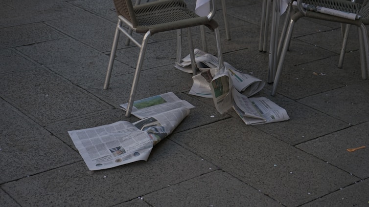 Newspapers are scattered on a dark pavement next to some metal-framed chairs. The scene appears to be outdoors in a public area, possibly a cafe or a street with seating.