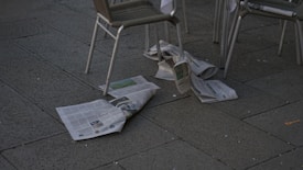 Newspapers are scattered on a dark pavement next to some metal-framed chairs. The scene appears to be outdoors in a public area, possibly a cafe or a street with seating.