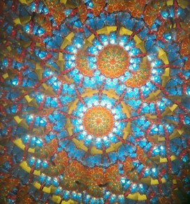 A vibrant kaleidoscopic pattern featuring intricate geometric shapes with a radial symmetry. The design incorporates bright colors such as blue, orange, red, and yellow, creating a visually striking mandala-like appearance.