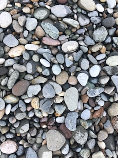 A collection of smooth, multicolored pebbles of various sizes and shapes tightly packed together. The stones range from gray to beige, with some showing hints of green, brown, and pink. The surface texture of the pebbles is generally smooth, with a few displaying small specks and patterns.