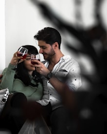 A couple is sitting closely together on a couch, holding and clinking glasses of red wine. They appear to be enjoying each other's company in a cozy, intimate setting.