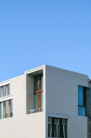 A modern building with clean lines and large windows, featuring a minimalist design. The structure has a mix of light-colored concrete and dark wood accents, set against a clear blue sky.