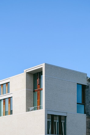 A modern building with clean lines and large windows, featuring a minimalist design. The structure has a mix of light-colored concrete and dark wood accents, set against a clear blue sky.