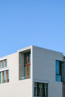 A modern building with clean lines and large windows, featuring a minimalist design. The structure has a mix of light-colored concrete and dark wood accents, set against a clear blue sky.