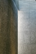 A minimalist architectural structure with textured concrete walls forming a corner. The surfaces are bathed in soft, natural light, casting subtle shadows that emphasize the raw material.