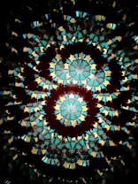 A vibrant and complex pattern of colorful stained glass resembling a kaleidoscope. The design features symmetrical, circular motifs with intricate geometric shapes in shades of red, blue, yellow, and green, radiating from the center.