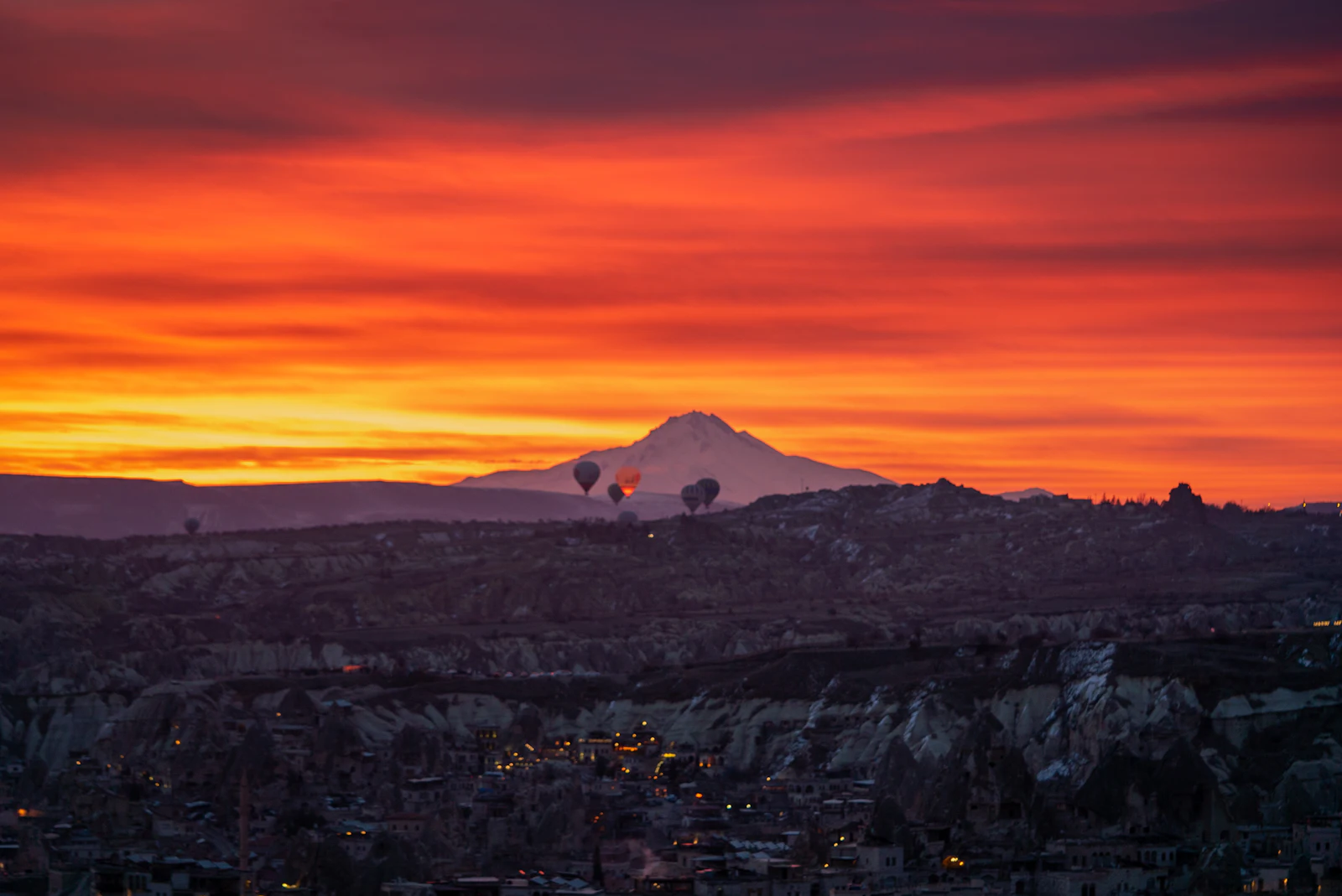 Hot air balloons drifting over Cappadocia's rock valleys at dawn under a fiery orange sky with Mount Erciyes behind