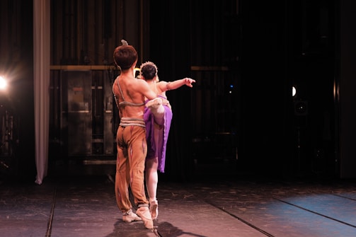 Two ballet dancers are on stage, captured from behind. The male dancer is wearing a form-fitting costume in shades of brown, and his arm is around the female dancer's waist. The female dancer is dressed in a purple outfit with a tutu, balanced on pointe shoes. They appear to be in a graceful pose with focus on their poised posture and elegance. Stage lights in the background create a dramatic ambiance with softer shadows.