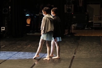 Two young dancers are standing on a dimly lit stage, dressed in ballet attire with light blue skirts and masks. The surrounding area appears to be a backstage setting with various equipment and cables visible in the shadows.