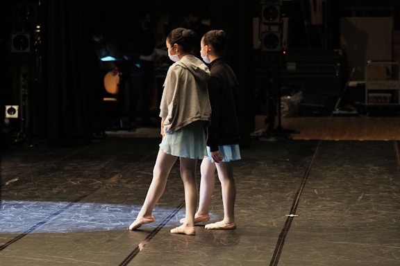 Two young dancers are standing on a dimly lit stage, dressed in ballet attire with light blue skirts and masks. The surrounding area appears to be a backstage setting with various equipment and cables visible in the shadows.