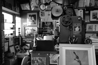 An old, vintage film projector is surrounded by various framed photographs and documents in a cluttered, cozy space. The room contains a mix of neatly stacked books, framed images, and office supplies, creating an atmosphere rich with history and nostalgia.