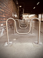 A row of metal bike racks is aligned under a covered area with a brick wall on the left. The perspective creates a tunnel-like effect, leading to a distant, light-filled opening at the end. The lighting is dim, suggesting it is indoors or under a structure.