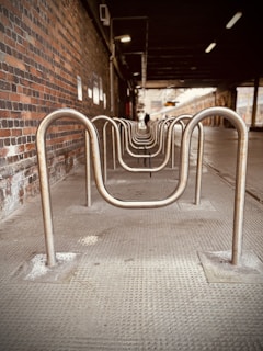 A row of metal bike racks is aligned under a covered area with a brick wall on the left. The perspective creates a tunnel-like effect, leading to a distant, light-filled opening at the end. The lighting is dim, suggesting it is indoors or under a structure.