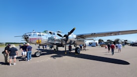 A vintage military aircraft with shiny metallic surfaces is parked on an airfield. The plane has the name 'Panchito' painted on its nose along with a cartoon image. Several people are gathered around the airplane, some observing and others taking photographs. The sky is clear and blue, adding to the bright atmosphere of the scene.