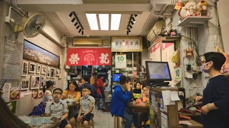 A bustling restaurant interior with several people sitting at tables. The walls display photographs and various decorations, including red banners with Chinese characters. A wall-mounted fan and an air conditioning unit are visible, and a person wearing a mask stands by a cash register. The atmosphere is lively and crowded with a mix of adults and children.