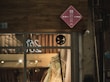A storefront of a clothing or bridal shop is seen, showcasing a white gown displayed behind a large glass window. Above the window, a red diamond-shaped sign with writing is mounted on a wood-textured exterior wall.