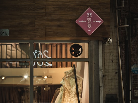 A storefront of a clothing or bridal shop is seen, showcasing a white gown displayed behind a large glass window. Above the window, a red diamond-shaped sign with writing is mounted on a wood-textured exterior wall.