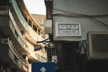 A street view featuring a narrow urban alleyway with tall buildings that have balconies with railings. There's a tiled wall with a bilingual street sign reading 'Calçada do Poço', and an air conditioning unit is visible. The lighting has a warm tone as it reflects off the building surfaces.