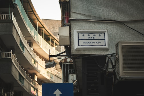 A street view featuring a narrow urban alleyway with tall buildings that have balconies with railings. There's a tiled wall with a bilingual street sign reading 'Calçada do Poço', and an air conditioning unit is visible. The lighting has a warm tone as it reflects off the building surfaces.