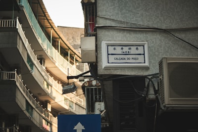 A street view featuring a narrow urban alleyway with tall buildings that have balconies with railings. There's a tiled wall with a bilingual street sign reading 'Calçada do Poço', and an air conditioning unit is visible. The lighting has a warm tone as it reflects off the building surfaces.