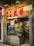 A cozy Asian restaurant storefront with prominent red signage and a glowing neon sign. The entrance includes a glass door, through which interior seating and customers are visible. Various promotional materials and Michelin stickers are displayed, indicating accolades and specialization in handcrafted soybean delicacies. The street sign reads 'Rua da Tercena'.
