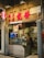 A cozy Asian restaurant storefront with prominent red signage and a glowing neon sign. The entrance includes a glass door, through which interior seating and customers are visible. Various promotional materials and Michelin stickers are displayed, indicating accolades and specialization in handcrafted soybean delicacies. The street sign reads 'Rua da Tercena'.
