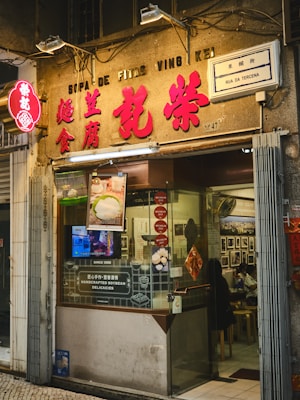 A cozy Asian restaurant storefront with prominent red signage and a glowing neon sign. The entrance includes a glass door, through which interior seating and customers are visible. Various promotional materials and Michelin stickers are displayed, indicating accolades and specialization in handcrafted soybean delicacies. The street sign reads 'Rua da Tercena'.