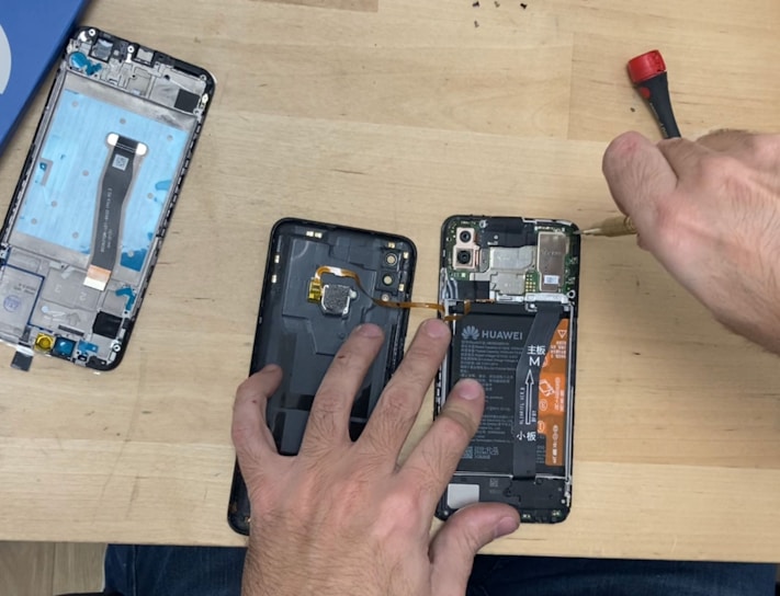 A person is disassembling a smartphone on a wooden table. The phone is partially opened, revealing internal components such as the battery and circuit boards. Tools like a screwdriver are visible, suggesting a repair or assembly process.