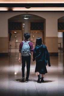 A couple is captured from the back as they walk hand in hand through a dimly lit, spacious hallway. The male is wearing a grey backpack and a maroon and grey jacket, while the female is dressed in black attire, holding her jacket over her shoulder. The hall features polished marble floors, tan walls, and a lit ceiling with a retro design. In the background, a staircase ascends through an arched doorway, adding depth to the setting.