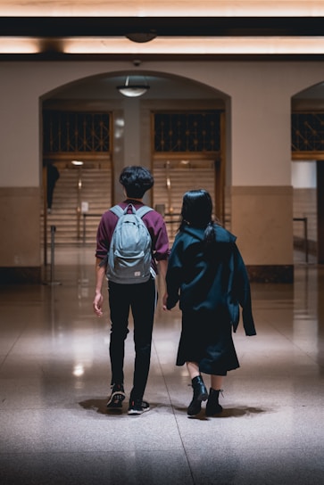 A couple is captured from the back as they walk hand in hand through a dimly lit, spacious hallway. The male is wearing a grey backpack and a maroon and grey jacket, while the female is dressed in black attire, holding her jacket over her shoulder. The hall features polished marble floors, tan walls, and a lit ceiling with a retro design. In the background, a staircase ascends through an arched doorway, adding depth to the setting.