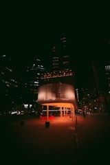 A modern cityscape at night features a cylindrical, illuminated pavilion with slatted walls. Red chairs and tables are arranged underneath, casting a warm glow on the surrounding pavement. Tall buildings in the background have scattered lights, contributing to the urban nighttime ambiance.
