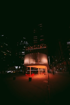 A modern cityscape at night features a cylindrical, illuminated pavilion with slatted walls. Red chairs and tables are arranged underneath, casting a warm glow on the surrounding pavement. Tall buildings in the background have scattered lights, contributing to the urban nighttime ambiance.