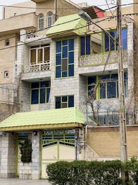 A multi-story residential building with a distinctive architectural style featuring green and blue accents. The structure includes large windows, a small balcony with intricate metal railings, and a front gate. The facade combines grey stonework with vibrant green and blue frames for the windows and roof elements.