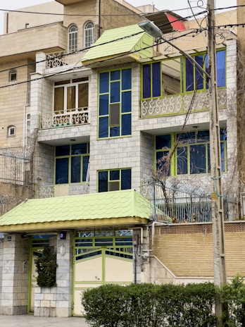 A multi-story residential building with a distinctive architectural style featuring green and blue accents. The structure includes large windows, a small balcony with intricate metal railings, and a front gate. The facade combines grey stonework with vibrant green and blue frames for the windows and roof elements.