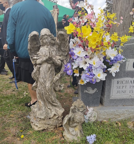 An ornate stone angel statue is positioned next to a gravestone adorned with a bouquet of colorful flowers, including yellow, white, and purple blooms. Several people in dark clothing are gathered in the background, suggesting a somber occasion.