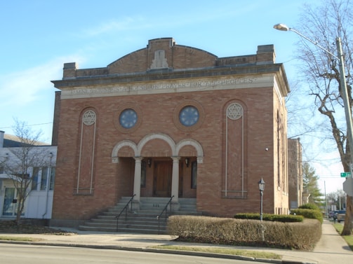 A brick building with arched entryway and steps leading up, featuring prominent Star of David symbols and an inscription above the entrance. The structure is flanked by bushes and trees, and sits on a corner with a street lamp and a visible street sign.